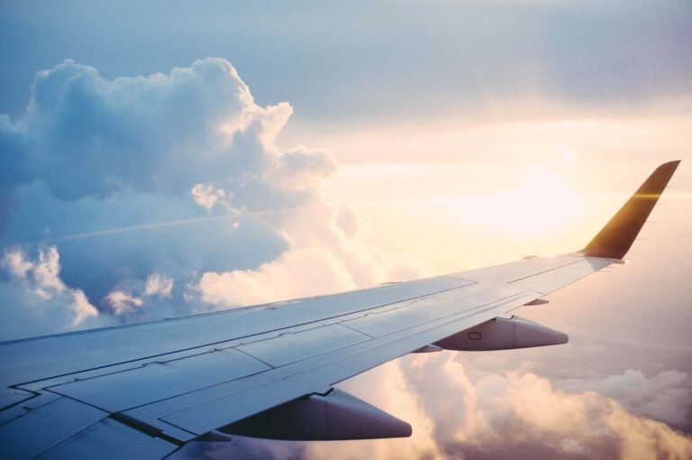 Ala de un avión vista desde una ventanilla, con nubes blancas y suaves que se destacan sobre un cielo azul y sereno al atardecer.