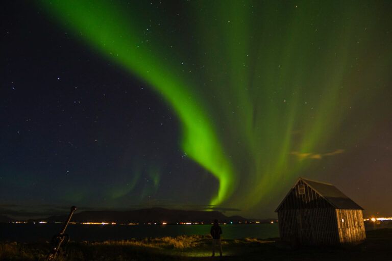 Aurora boreal verde sobre el horizonte y una pequeña cabaña de madera en la costa de Islandia.