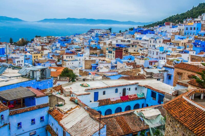 Vista panorámica de los icónicos edificios pintados de azul en la medina de Chefchaouen, con montañas al fondo bajo un cielo nublado.