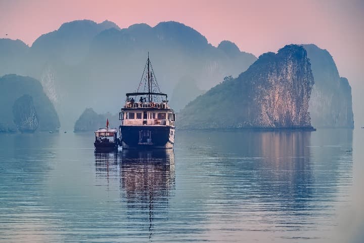 barco grande y pequeño navegando en ha long bay, detrás formaciones rocosas y cielo color rosado - weroad