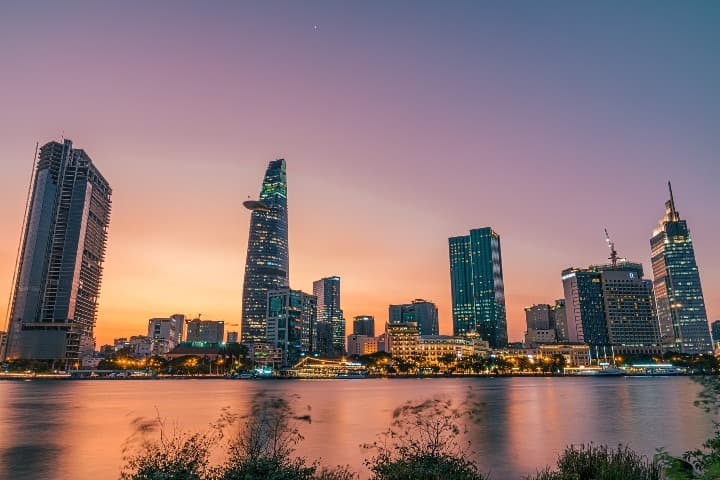 ho chi minh city vista desde el borde de rio al atardecer, rascacielos y edificios iluminados durante el atardecer rosado - weroad