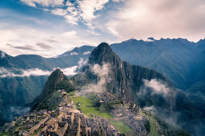 Vista panorámica de la ciudadela inca de Machu Picchu, mostrando las ruinas, terrazas verdes y el icónico pico de Huayna Picchu al fondo, rodeado de montañas andinas y nubes bajas.