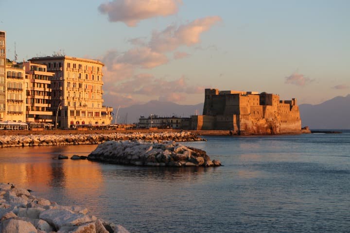 castel dell'ovo en medio al mar, al fondo edificios iluminados por la luz del atardecer - weroad