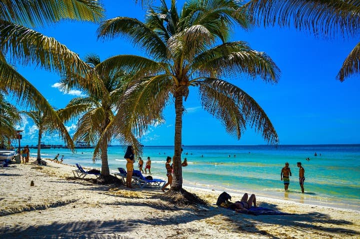 Playa paradisíaca en Cuba con palmeras, arena blanca y aguas turquesas bajo un cielo azul brillante con bañistas.