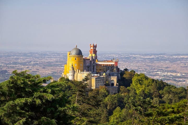 edificio amarillo y rojo, el palacio nacional de pena en sintra encima de un promontorio con vegetación - weroad