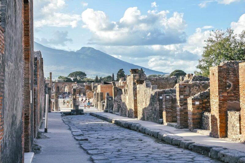 calles adoquinadas de pompeya con restos de las casas, al fondo el vesubio y cielo con nubes - weroad