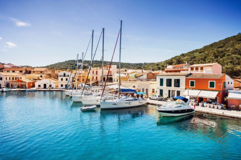 barcos de vela en el puerto de paxos, al fondo casitas, montaña y cielo azul - weroad