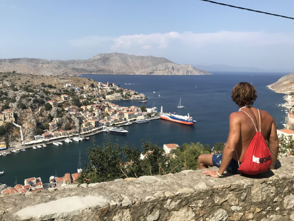 coordinador weroad de espaldas, con mochila roja, sentado en un murillo en la isla de symi, cerca de rodas, mirando casitas y mar