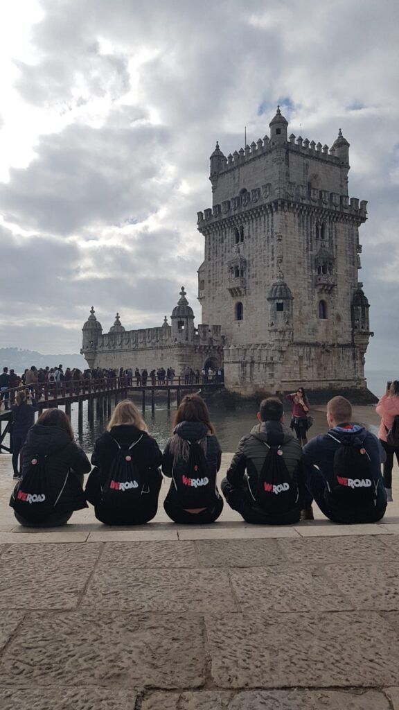 5 chicos sentados de espaldas delante de la torre de belem, en lisboa, tienen mochilas negras de weroad