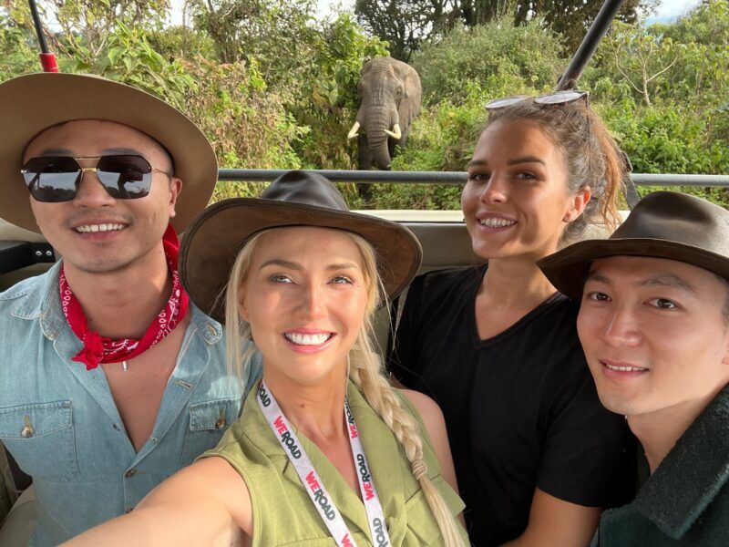 selfie de cuatro vajeros weroad durante un safari en tanzania, en un coche abierto y con detrás la cara de un elefante