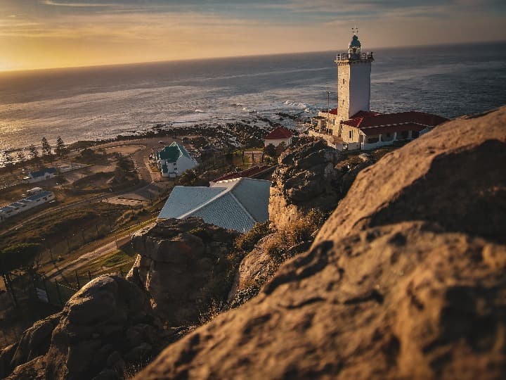 casa con faro y al fondo el mar, en primer plano una roca en mossel bay, en la garden route de sudáfrica