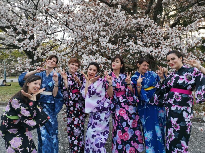 Un grupo de viajeras sonriendo y haciendo el signo de la paz con kimonos tradicionales frente a cerezos en flor en Japón.