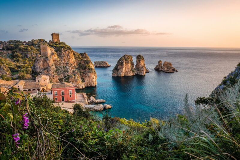 Vista panorámica de la Tonnara di Scopello al atardecer, con sus farallones rocosos emergiendo del mar cristalino en Sicilia.