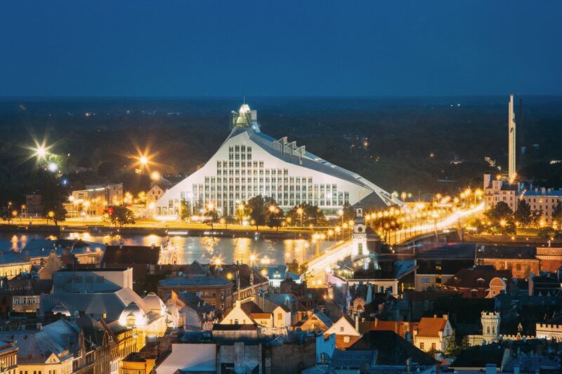 Vista nocturna iluminada de la Biblioteca Nacional de Letonia y el skyline de Riga reflejado en el río Daugava.