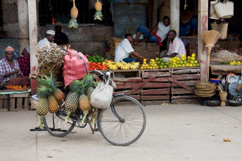 Una bicicleta cargada de piñas estacionada frente a un concurrido mercado de frutas y verduras en Zanzíbar.