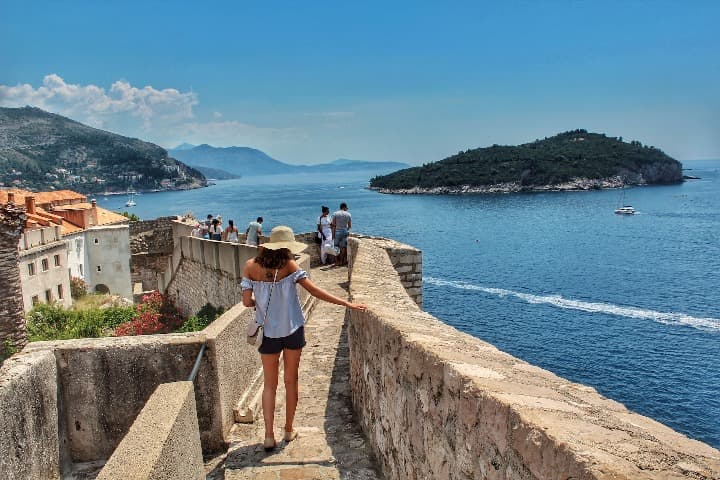 murallas de la ciudad de dubrovnik, algo que ver en croacia, una chica camina de espaldas, al fondo mar y una isla - weroad