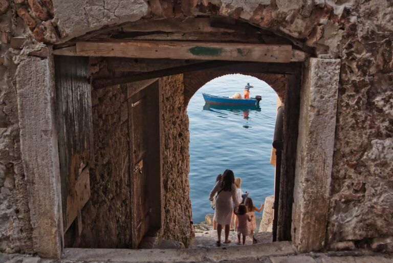 puerta de edificio a través de la cual se ve mar, barquito y personas llegando a la playa de rovinj, croacia - weroad