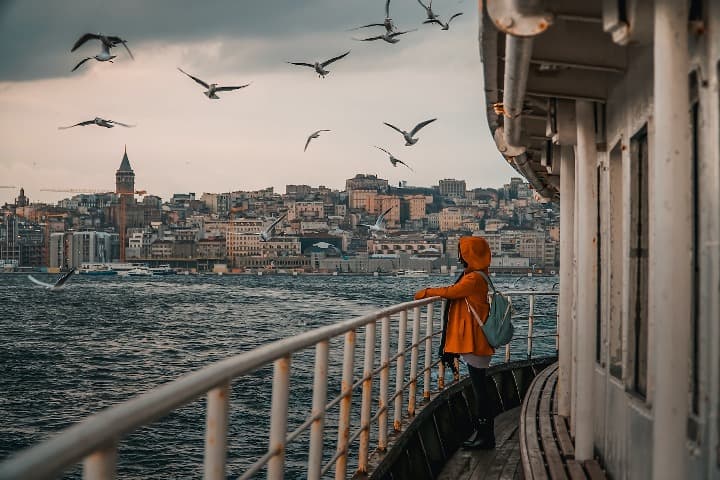 mujer con impermeable naranja encima de un barco, al fondo edificios de la ciudad de estambul y encima pajaros volando