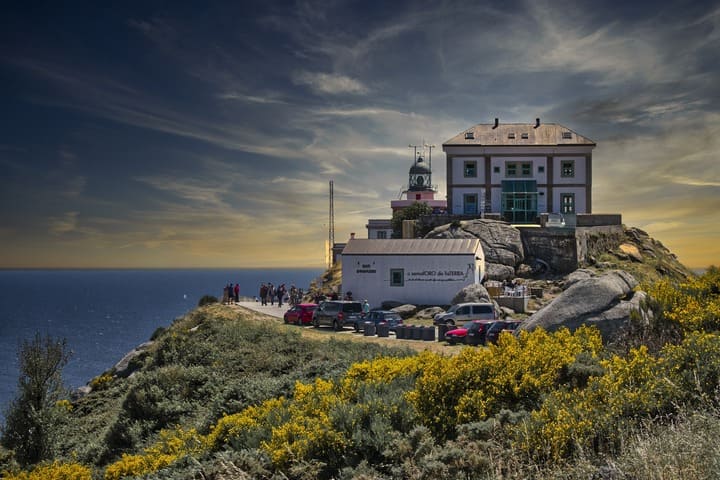 edificio en la cumbre de una montaña con hierba y flores delante, en finisterre, galicia - weroad