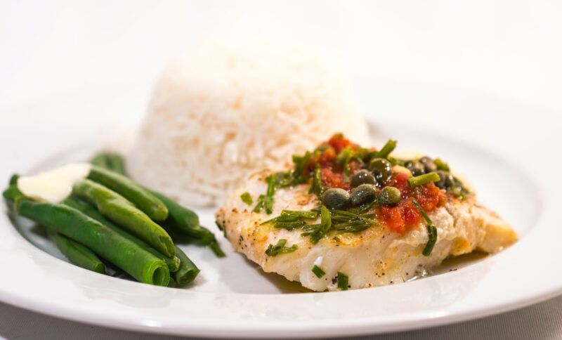 Un lomo de bacalao a la plancha con salsa de tomate y alcaparras, servido con arroz blanco y judías verdes en un plato blanco.