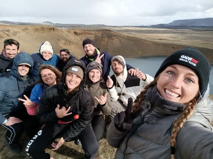 Viajeros de WeRoad sonríe durante un selfie grupal frente a un lago en un paisaje volcánico de Islandia.