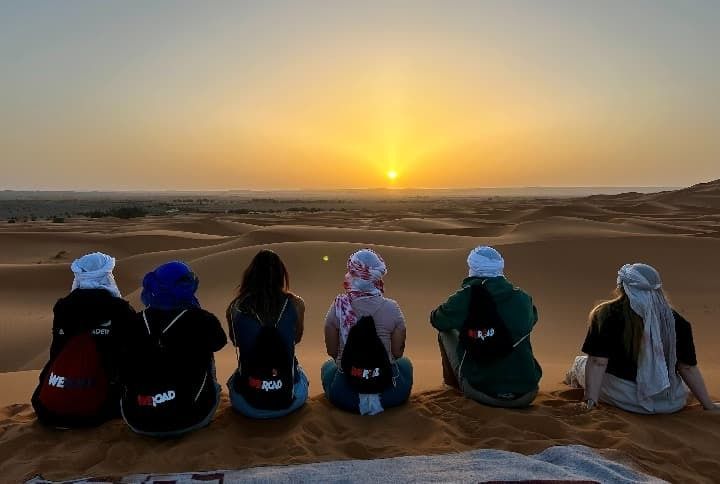 Viajeros sentados en una duna, contemplando la puesta de sol sobre el desierto en Marruecos.