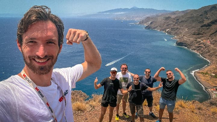 selfie de chico con camiseta weroad y otros viajeros detrás, de fondo el mar de las canarias, destino ideal para el puente de todos los santos