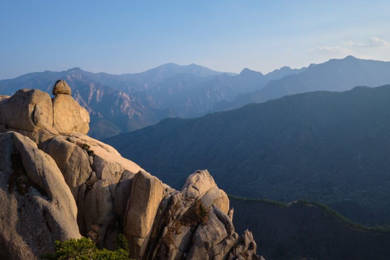 Vista panorámica desde el pico Ulsanbawi al atardecer, mostrando formaciones rocosas escarpadas y capas de montañas azuladas bajo un cielo despejado en el Parque Nacional Seoraksan.