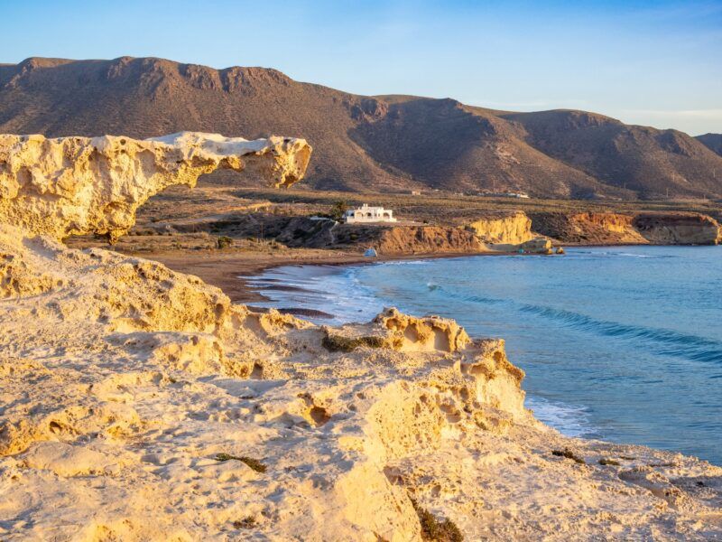 Formaciones rocosas de dunas fósiles en la playa de Los Escullos, Cabo de Gata, con el mar azul y montañas al fondo.