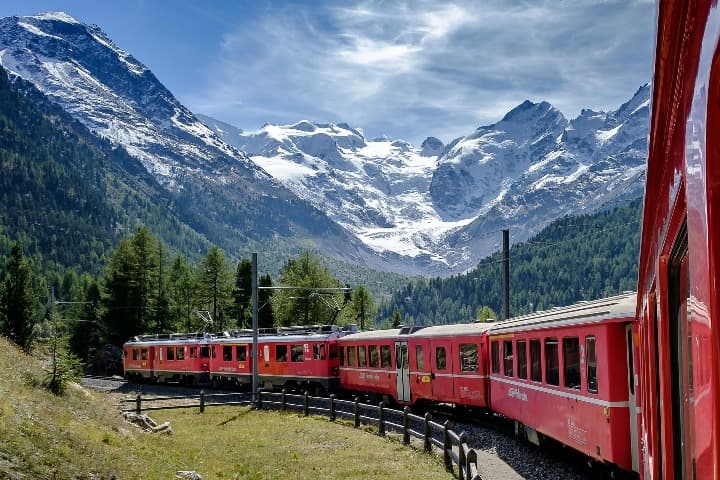 tren blanco y rojo en un paisaje de montañas y árboles en suiza, bernina express, uno de los mejores viajes en tren en europa