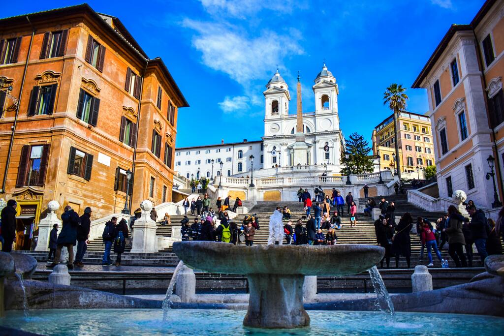 Vista panorámica de la Plaza de España en Roma, mostrando la emblemática escalinata de Trinità dei Monti y la fuente de la Barcaccia en un día soleado y animado.