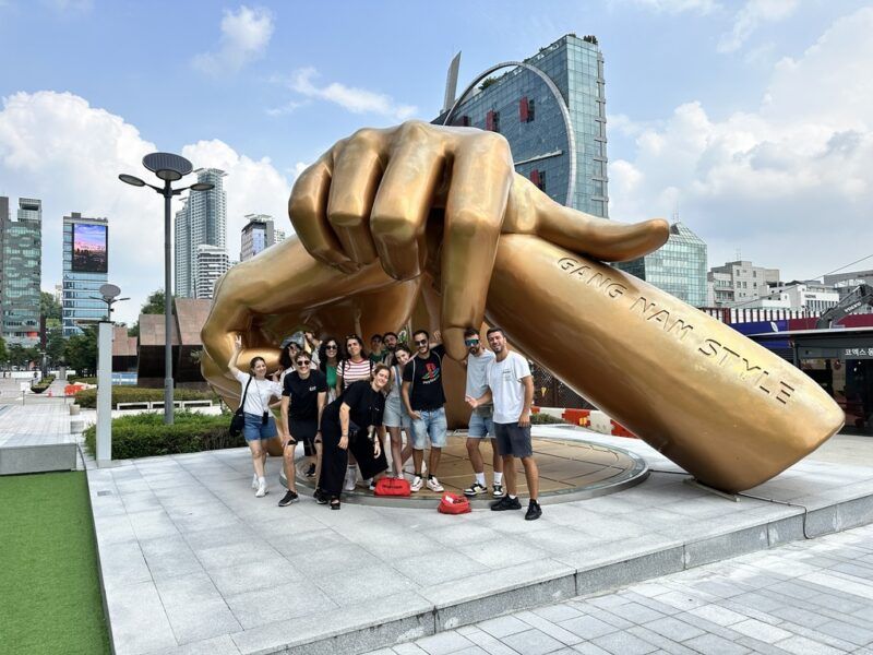 Un grupo de viajeros WeRoad posa frente a la gran escultura dorada de manos 'Gangnam Style' en el distrito de Gangnam, Seúl, rodeados de edificios modernos.