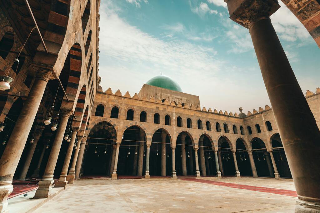 Vista interior de una mezquita histórica en El Cairo con columnas y una cúpula verde.
