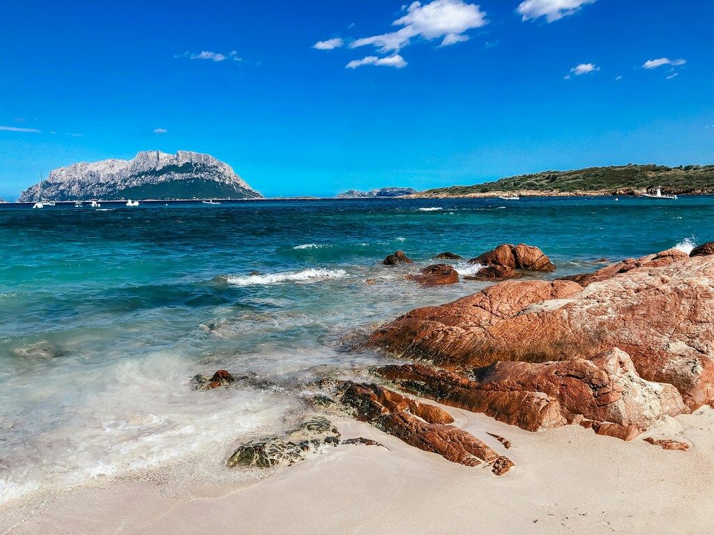 Vista de la playa de arena blanca con rocas rojizas y aguas cristalinas en Cerdeña, con la isla de Tavolara al fondo