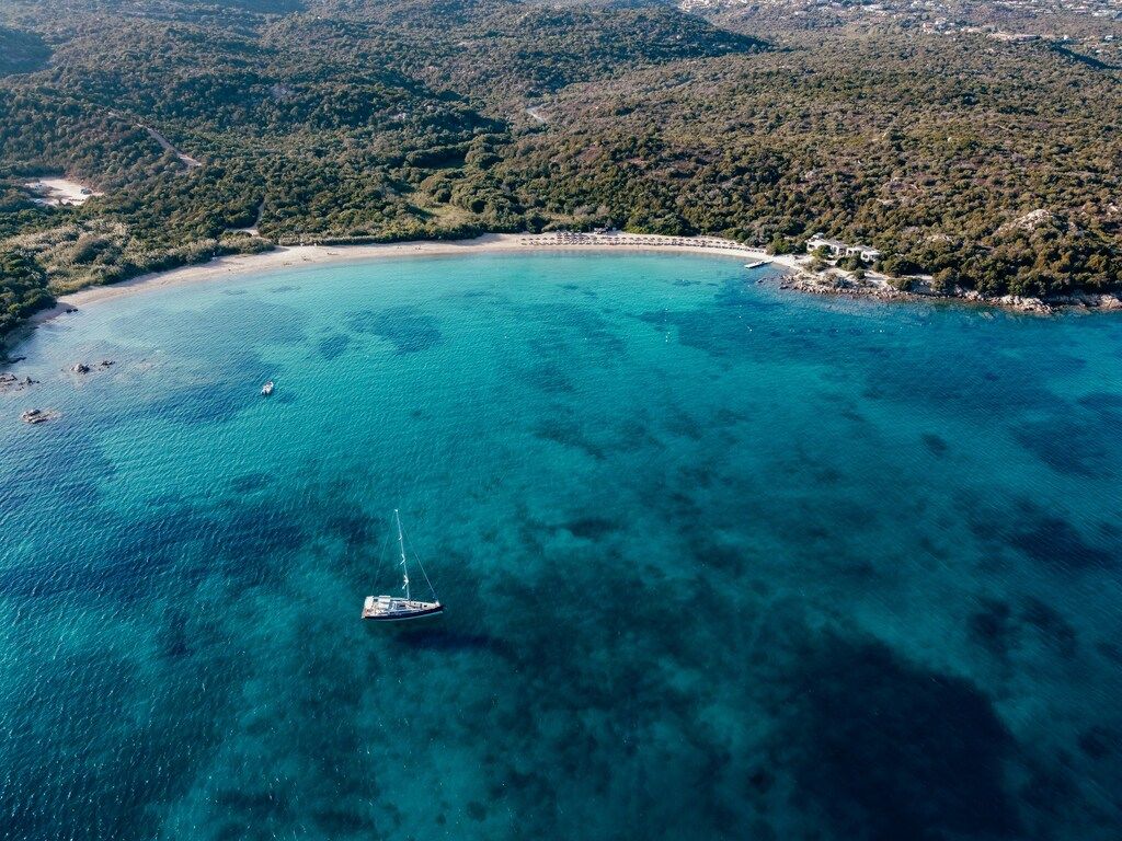 Vista aérea de una playa en Cerdeña con una barca de vela sobre aguas cristalinas y vegetación mediterránea alrededor
