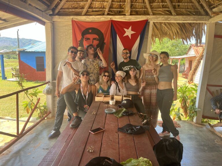 Un grupo de viajeros de WeRoad posando con cigarros frente a una bandera cubana y un retrato del Che Guevara en una cabaña al aire libre.