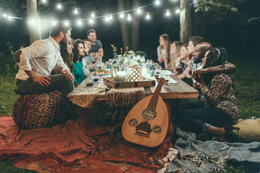 Cena al aire libre con luces colgantes, donde un grupo de personas comparte una comida en un ambiente acogedor y multicultural.