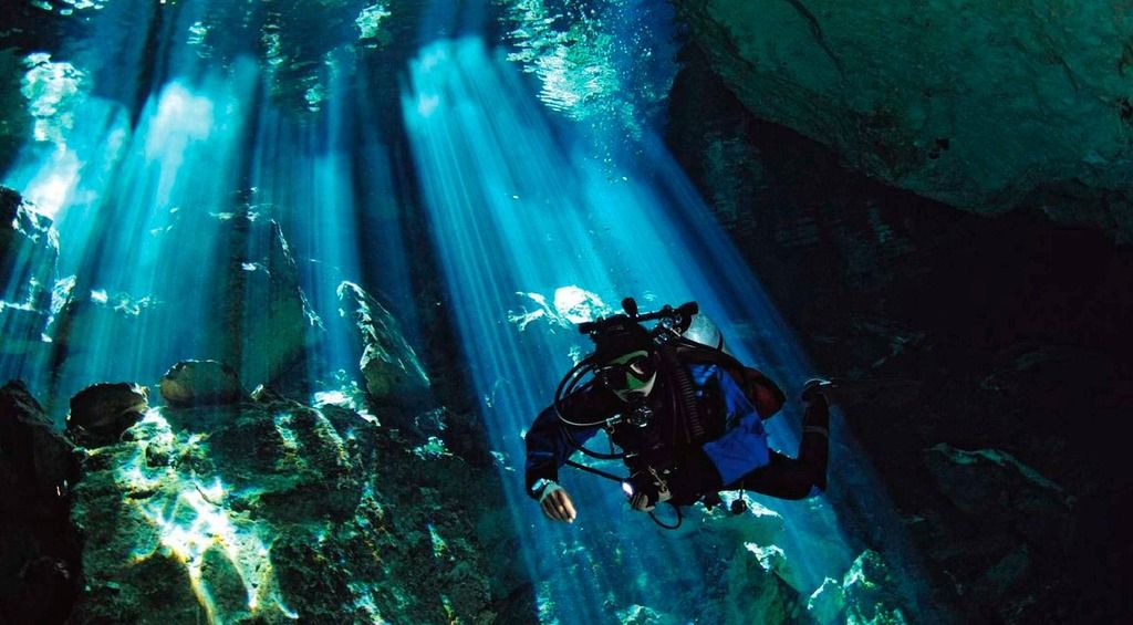 Buceador explorando un cenote subterráneo con rayos de luz filtrándose entre las rocas.