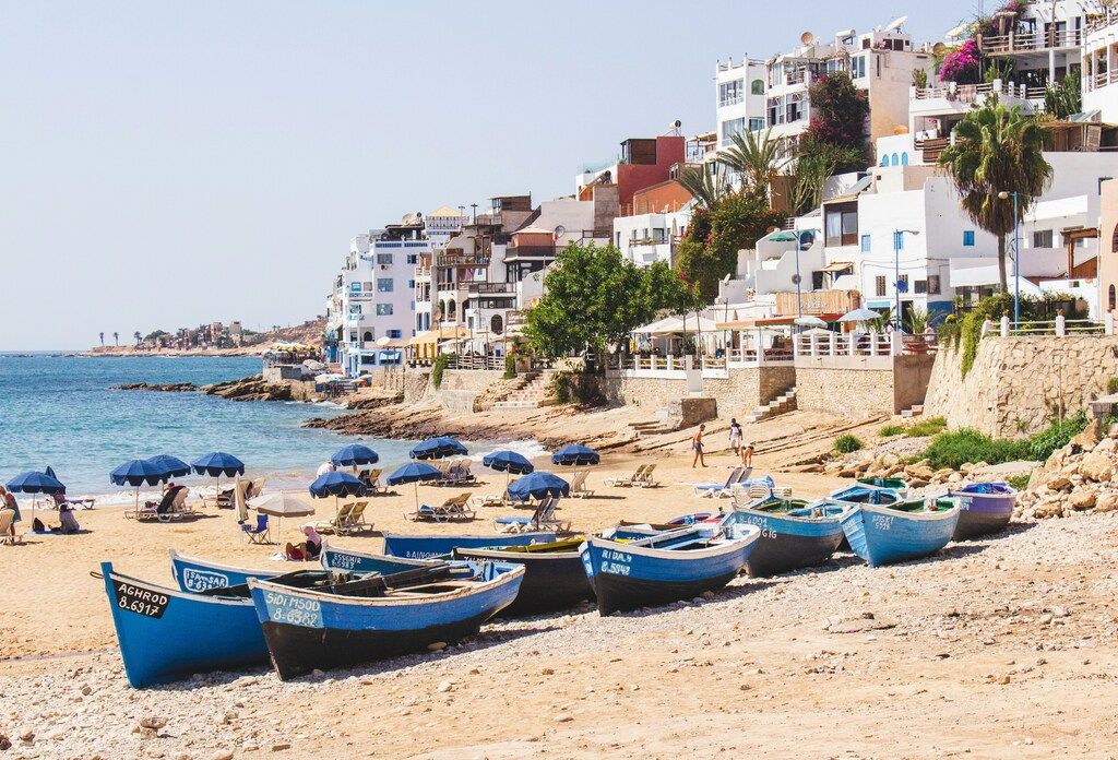 Pequeñas barcas azules sobre la arena en una playa marroquí con casas blancas frente al mar.