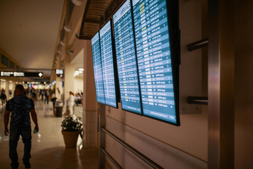 Pantallas digitales con horarios de llegadas en un aeropuerto concurrido, con pasajeros caminando por el pasillo.