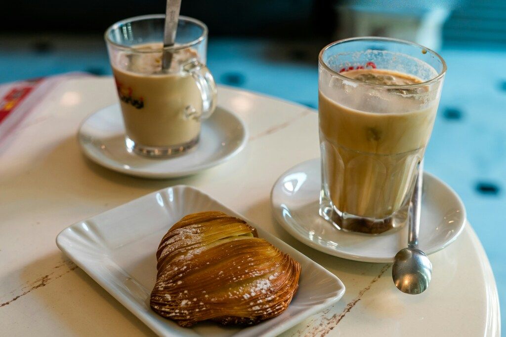 Una sfogliatella riccia espolvoreada con azúcar glas en un plato blanco, junto a dos vasos de café con leche fría y cucharas, en una mesa de mármol.