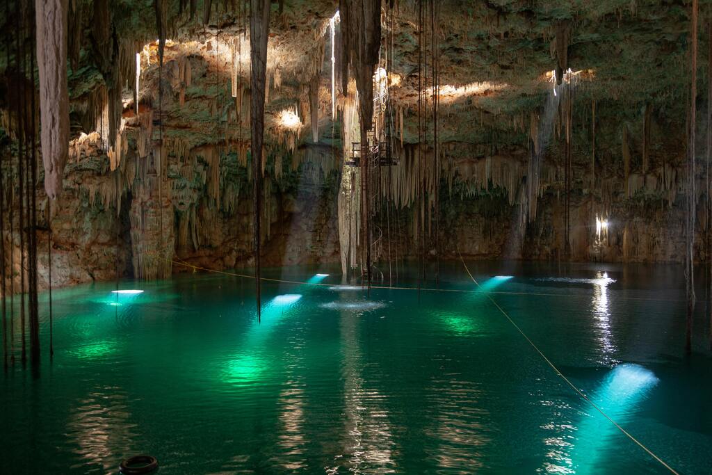 Interior de un cenote iluminado con luces de colores, mostrando estalactitas y agua verde esmeralda.