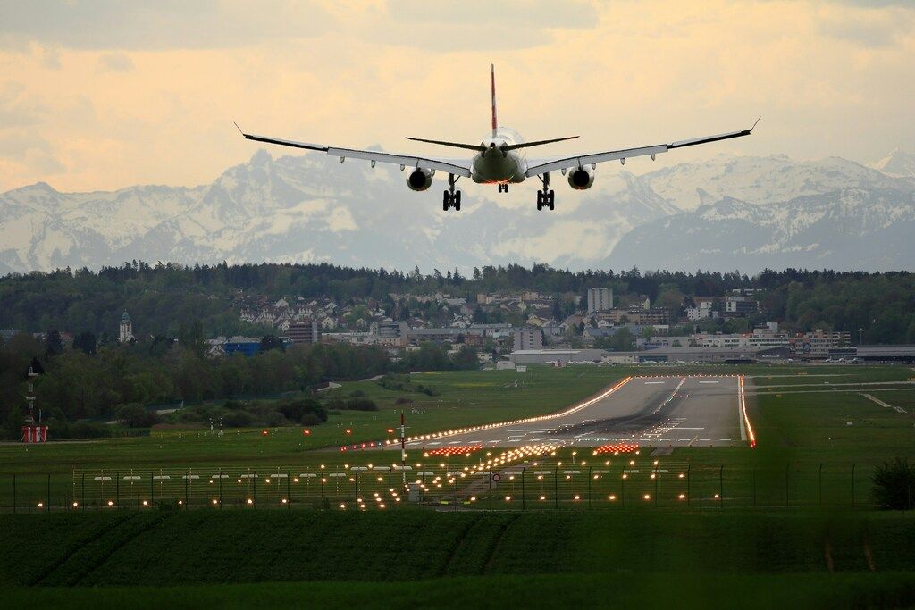 Avión aterrizando en una pista iluminada con vista a una ciudad y montañas nevadas al fondo.