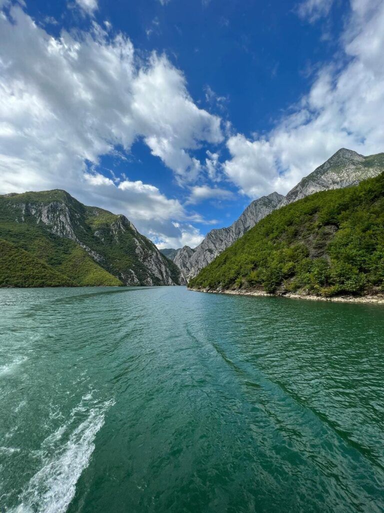 Paisaje del lago Koman visto desde una embarcación, con aguas verdes, montañas escarpadas y un cielo con nubes dramáticas.