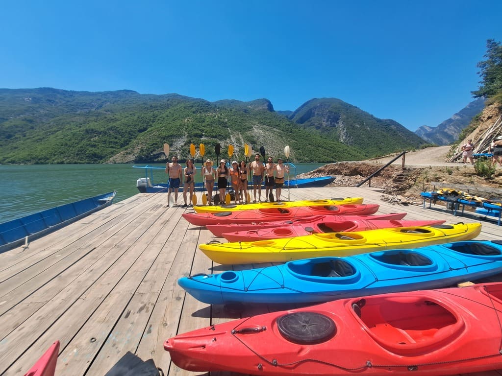 Grupo de kayaks rojos, amarillos y azules alineados en un muelle de madera junto al lago Koman, con colinas verdes al fondo.