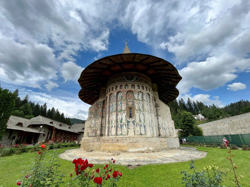 Iglesia ortodoxa con frescos exteriores en un jardín con flores rojas, bajo un cielo parcialmente nublado.