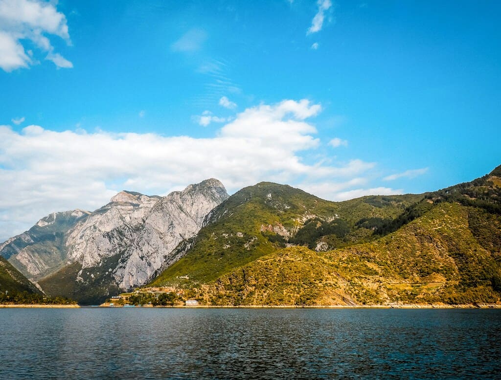 Vista panorámica del lago Koman rodeado de montañas verdes y escarpadas bajo un cielo parcialmente nublado en Albania.