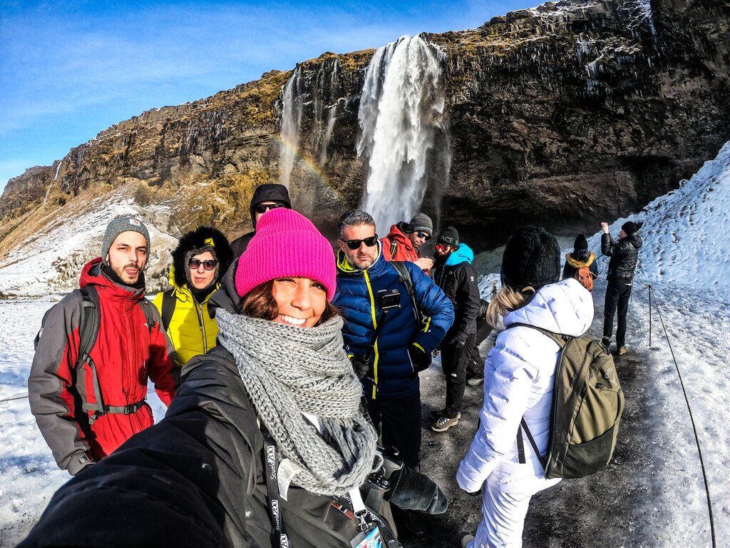 Grupo de viajeros WeRoad posando frente a la cascada Seljalandsfoss, rodeada de nieve y con un arco iris en el fondo en Islandia.