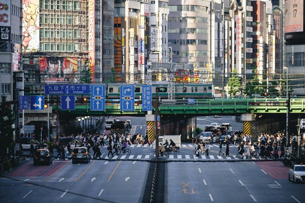 Multitud cruzando en una gran intersección con tren pasando por el puente verde.