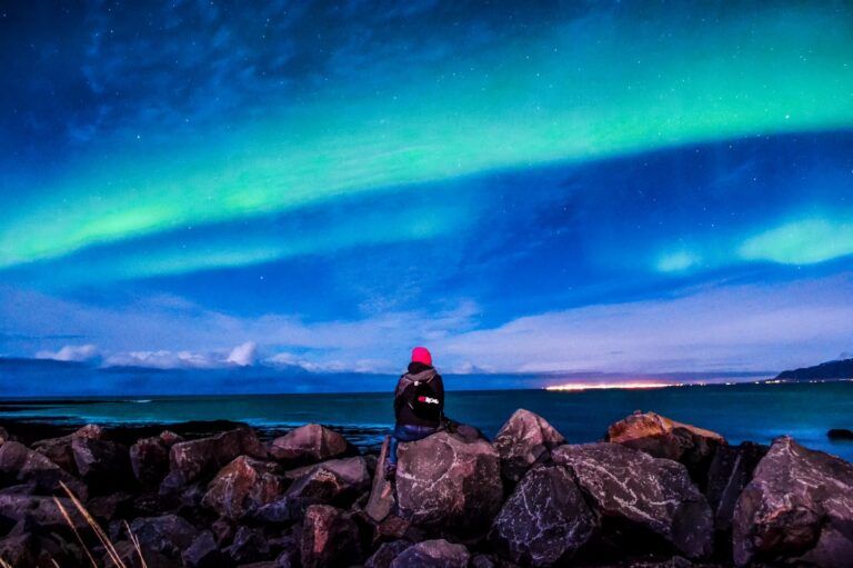 Persona sentada en rocas mirando la aurora boreal sobre el mar en Islandia.